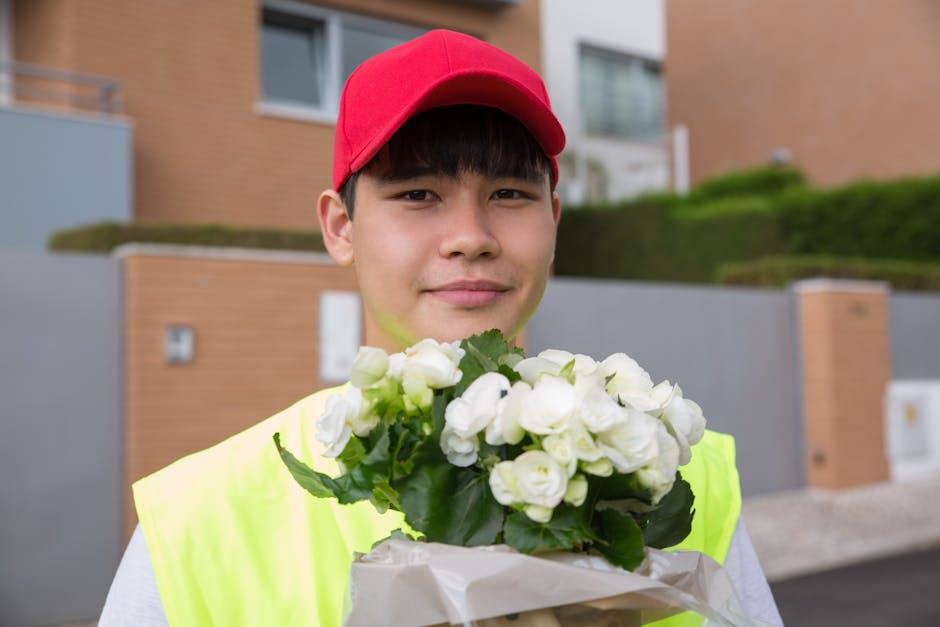 A young delivery person with light skin, wearing a bright yellow vest and a red cap, is holding a bouquet of fresh white flowers wrapped in brown paper. The bouquet consists of numerous small, tightly