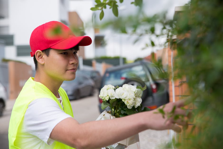 A young male florist wearing a red cap and a high-visibility yellow vest is arranging a bouquet of fresh white roses, held in a clear plastic wrapper, outdoors near a brick wall and parked cars. The b