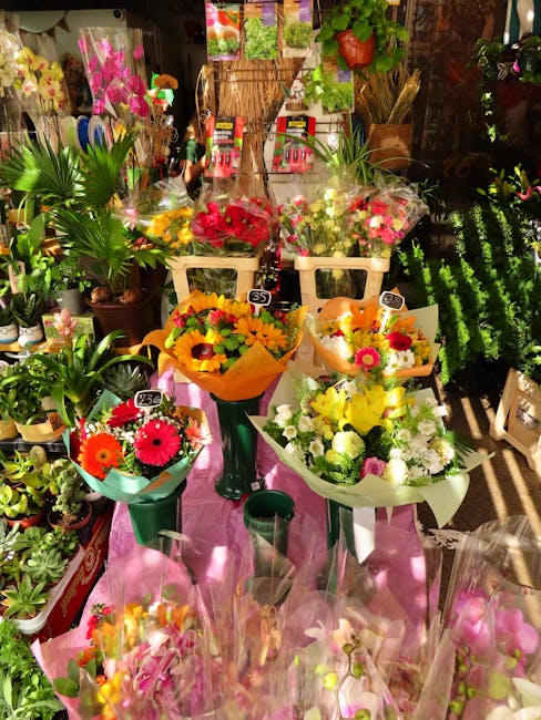 A vibrant display of fresh floral arrangements and bouquets arranged on a pink table within a flower shop. The flowers include bright pink, yellow, orange, and white blooms such as gerbera daisies, ch