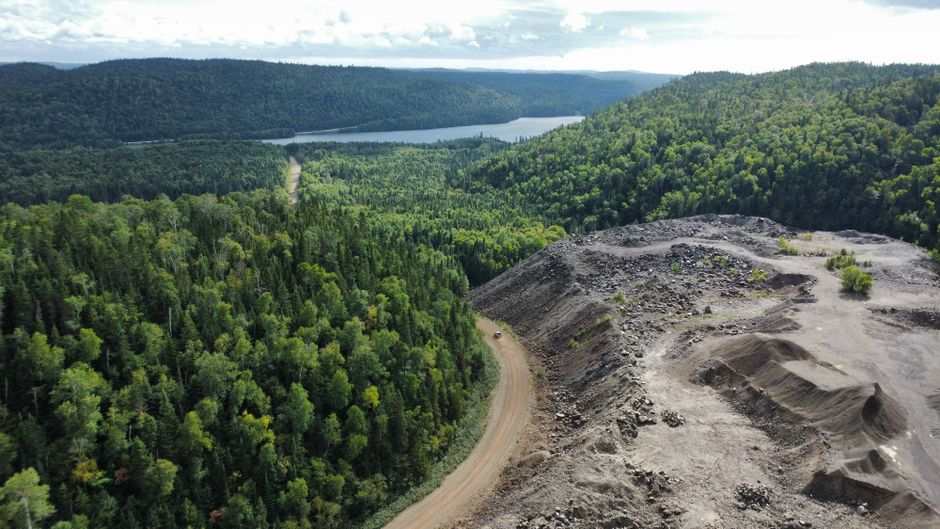 An aerial view showing dense green woodland with a narrow dirt road winding through the trees, leading towards a lake in the distance. To the right, there is a cleared area with loose soil, gravel, an