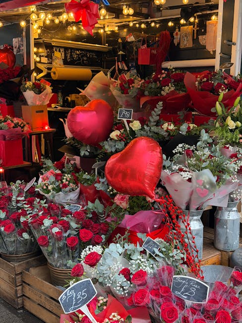 A vibrant floral display at a florist shop featuring an assortment of fresh red and white flowers, including roses and carnations, arranged in bouquets and wrapped in pink, red, and white paper. Two l